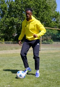 Atleta in una vivace felpa gialla e pantaloni neri, che palleggia un pallone da soccer sull'erba. Pallone blu e bianco con dettagli a fantasia ben visibili.
