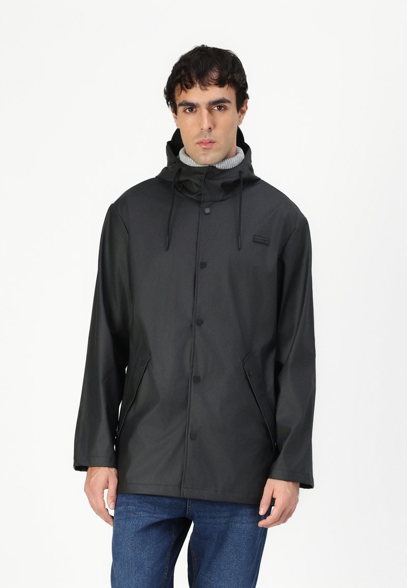 Veste imperméable noire avec capuche, boutons-pression, poches latérales et cordons de serrage réglables. Portee par-dessus un pull gris clair et un jean.