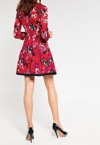 Robe fleurie rouge à manches longues, accents noirs à l'ourlet, jupe évasée, taille cintrée, associée à des chaussures à talons hauts noires.