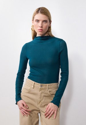 Teal ribbestrikket turtleneck med volangkrage, lagt sammen med beige høyt liv bukser. Stoffet ser mykt ut med en tettsittende silhuett.
