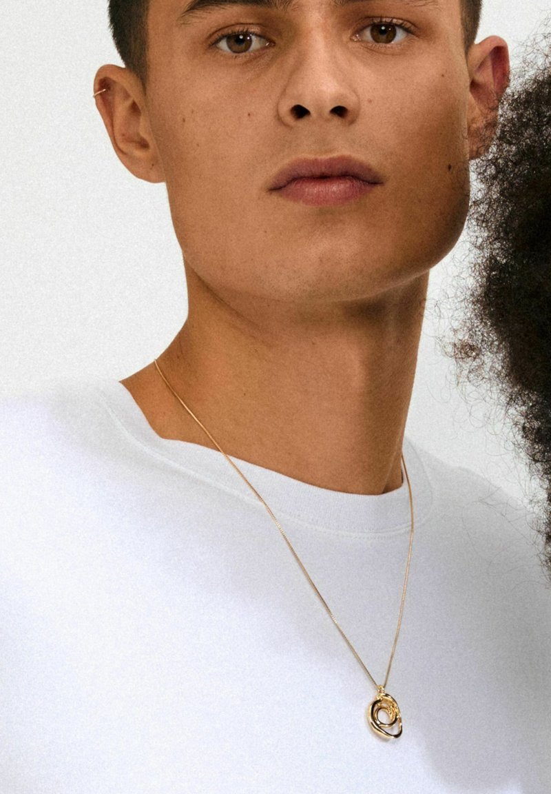 Halskæde med guld tone og sammenflettet cirkulært design, båret over en enkel hvid t-shirt. Glat metaloverflade, fin kæde.
