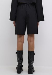 Pantalones cortos negros a medida, de longitud a mitad del muslo, con cintura elástica y bolsillos laterales. Llevados con botas de cuero negro hasta la rodilla.