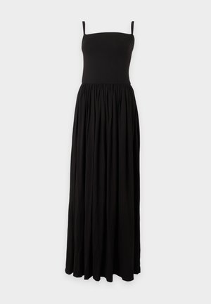 Femme aux longs cheveux foncés portant une robe maxi noire sans manches, debout pieds nus sur un fond clair uni.