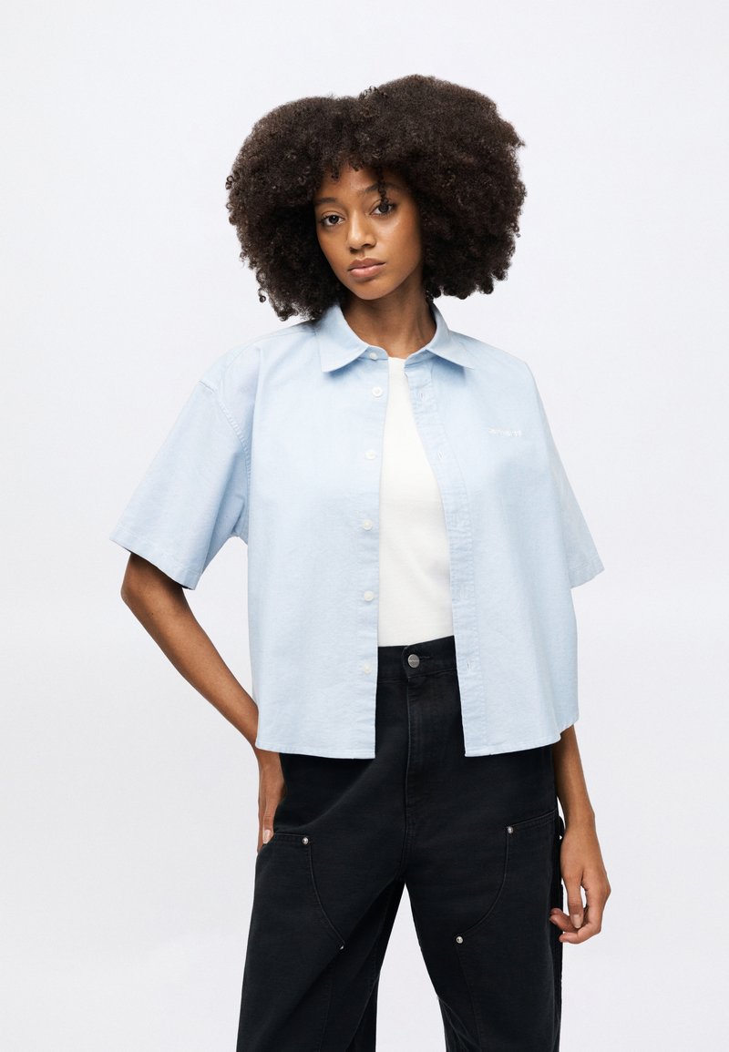 Chemise bleu clair à manches courtes en coton, avec une coupe décontractée et des détails de broderie blanche, associée à un pantalon noir taille haute.