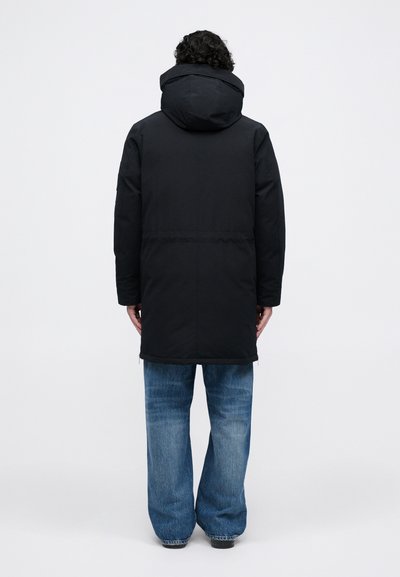 Juodas gobtuvą turintis parkas su ilga forma, pasižymintis lygiu paviršiumi, elastinėmis rankovėmis ir susiauču liemeniu. Dėvimas virš šviesiai mėlynų plačių džinsų.