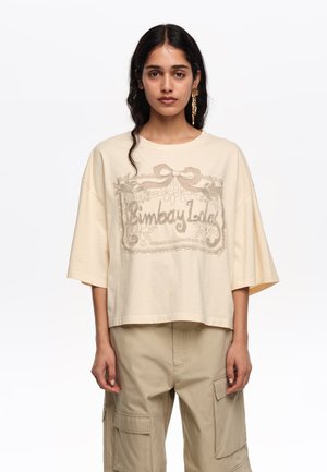 Mlada ženska z dolgimi temnimi lasmi nosi preveliko bež vezeno majico z napisom "Bimba y Lola" in ujemajoče se cargo hlače, nevtralna ozadje.