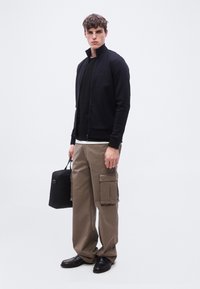 Νεαρός άνδρας με μαύρο μπουφάν με φερμουάρ, μαύρο πουκάμισο, μπεζ παντελόνι cargo, μαύρα παπούτσια, κρατώντας μαύρη τσάντα σε απλό φόντο.