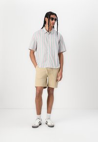 Chemise pour homme à manches courtes avec des rayures pastel (rose et menthe) assortie à un short beige. Lunettes de soleil noires et baskets blanches avec des accents noirs.