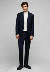 Marinefarbener strukturierter Blazer über einem weißen Rollkragenpullover, kombiniert mit maßgeschneiderten dunklen Hosen und schwarzen Schnürschuhen. Schlichtes, modernes Design.