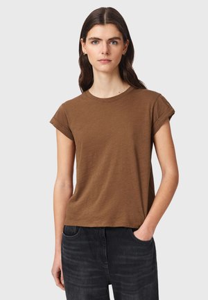 Junge Frau mit langen dunklen Haaren, die ein braunes kurzärmliges T-Shirt und dunkle Jeans trägt, mit einer Hand in der Tasche vor einem einfarbigen Hintergrund.