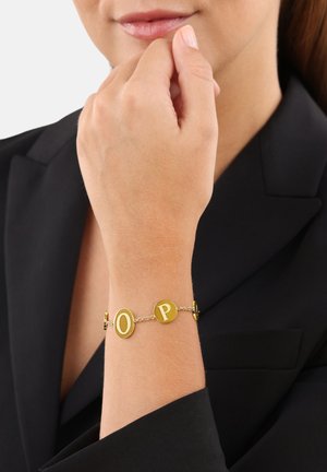 Pulsera de oro que presenta letras redondas "O" y "P", unidas por una delicada cadena. Superficie lisa y pulida con un acabado brillante.
