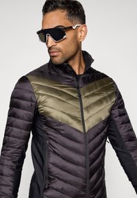 Siltumizolēts mētelis ar melnu un olīvu viļņotu rakstu, kas apvieno rāvējslēdzēju un moderna dizaina. Leņķainas melnas saulesbrilles ar atstarojošiem stikliem.