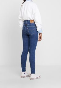 Vaqueros ajustados de denim en color azul, con una cintura alta, bolsillos traseros con costura de marca, y combinados con un sudadera blanca y zapatillas.
