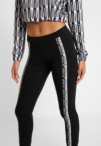 Donna che indossa leggings neri con strisce laterali bianche con il logo Adidas e un crop top nero e bianco della Adidas a maniche lunghe.