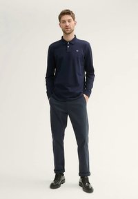 Temno modra polo majica z dolgimi rokavi in klasičnim ovratnikom z detajlom logotipa, kombinirana s temnimi hlačami in črnimi čevlji z vezalkami. Enobarvna, minimalističen dizajn.