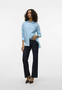 Light blue, long-sleeve button-up shirt made of textured fabric, paired with dark blue flared jeans and brown heeled shoes.
