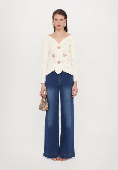 Knitted ivory sweater with a V-neck, decorative cutouts, and textured detailing, paired with high-waisted blue wide-leg jeans and a patterned bag.