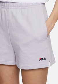 Pantaloni corti in cotone lilla con una cintura elastica, tasche laterali e un logo Fila in rosso, bianco e blu sull'orlo inferiore.