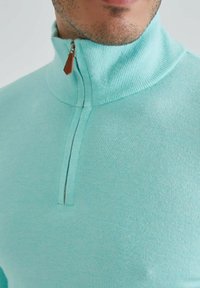 Pull léger bleu en tissu doux, avec un col à demi-zip et un tirant de fermeture Éclair en cuir marron, ainsi que des détails en côtes.