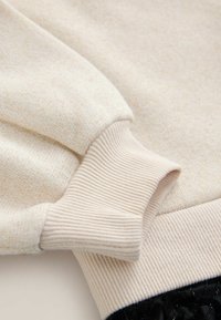 Cremfarbener Strickpullover mit einem gerippten Bündchen, das auf einem gerippten Taillenbund ruht, und eine weiche Textur sowie einen subtilen Schimmer im Stoff zeigt.