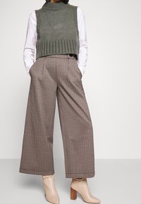 Olivengrüner gestrickter ärmelloser Pullover über einem weißen Hemd getragen, kombiniert mit braun karierten weit geschnittenen Hosen und nuden High Heels.