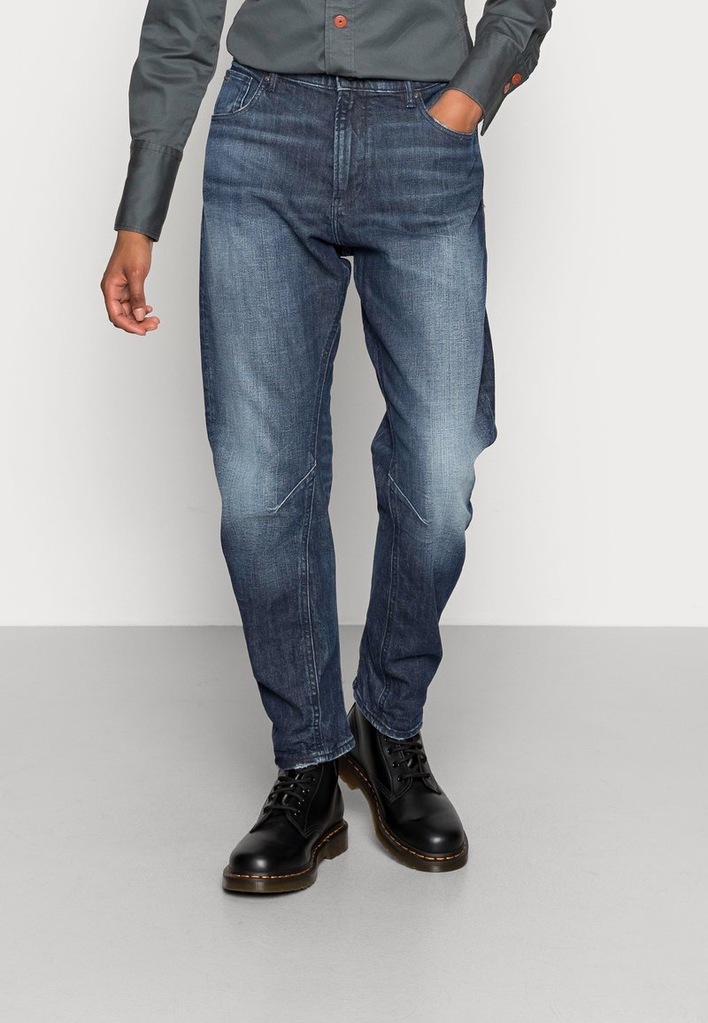 Dunkelblaue Jeans im Slim-Fit-Stil mit Fronttaschen und leicht verwaschenem Look, kombiniert mit schwarzen Stiefeln.