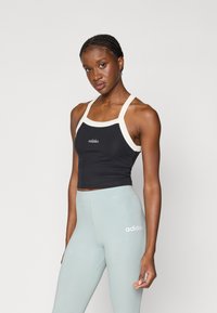 Juodas trumpas viršus su kreminėmis petnešėlėmis, turintis prigludusį dizainą ir siuvinėtą logotipą. Šviesiai mėlynos aukšto liemens legginsai su subtiliais ženklais.