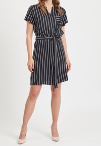 Vestido a rayas en blanco y negro con mangas cortas, cierre de botones en la parte delantera, bolsillo y un cinturón a la cintura. Tela suave, corte a la altura de la rodilla.