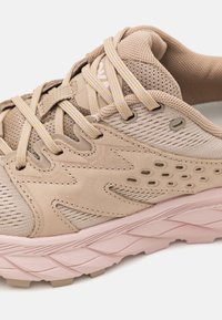 Lys beige løpesko med mesh-overdel, teksturert design med perforeringer, rosa mellomsåle og kontrasterende gul søm på snøringen.