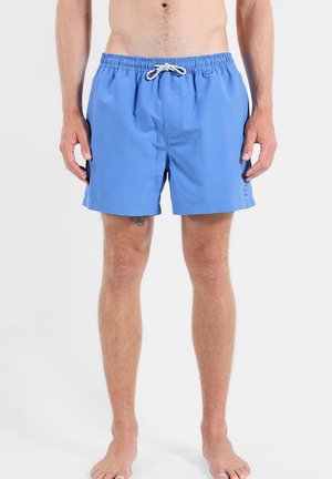 Homme portant un short de bain bleu clair avec un cordon blanc, debout pieds nus sur un fond blanc uni.