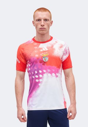 Joven con el pelo corto y rojo que lleva una camiseta deportiva multicolor en rosa, rojo y blanco con un emblema de pájaro en el pecho y pantalones cortos azul marino.