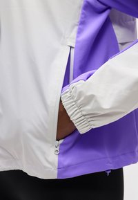 Χέρι μερικώς μέσα στην τσέπη με φερμουάρ ενός ανοιχτόχρωμου γκρι και μοβ μπουφάν με ελαστική μανσέτα στο μανίκι.