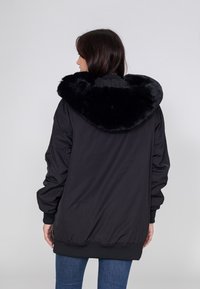 Zwarte bomberjack met een grote nepbont kraag, voorzien van een ritssluiting aan de voorkant en geribde manchetten. Achteraanzicht, gedragen met donkere jeans.