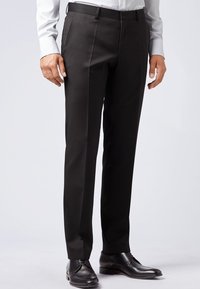 Formella svarta byxor bärs med svarta läderskor och en vit långärmad skjorta med knappar mot en enfärgad bakgrund.