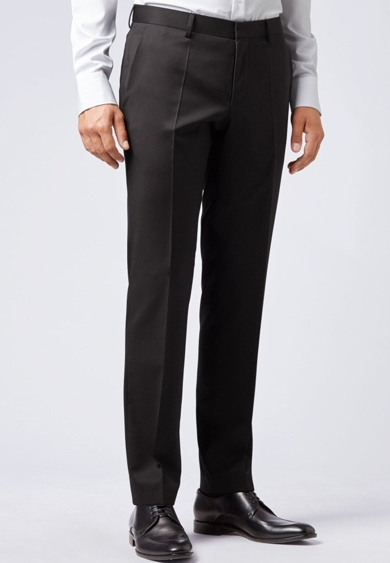 Formella svarta byxor bärs med svarta läderskor och en vit långärmad skjorta med knappar mot en enfärgad bakgrund.