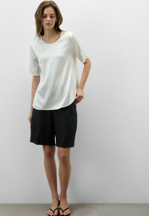 Frau trägt eine weiße kurzärmelige Bluse, schwarze knielange Shorts und schwarze Sandalen und steht vor einer schlichten weißen Wand.