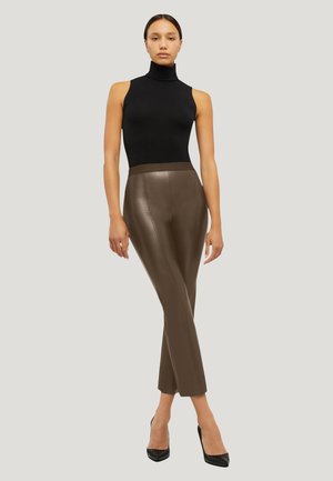 Donna con top nero senza maniche a collo alto, pantaloni aderenti in pelle marrone e tacchi alti neri, in piedi davanti a uno sfondo chiaro e semplice.