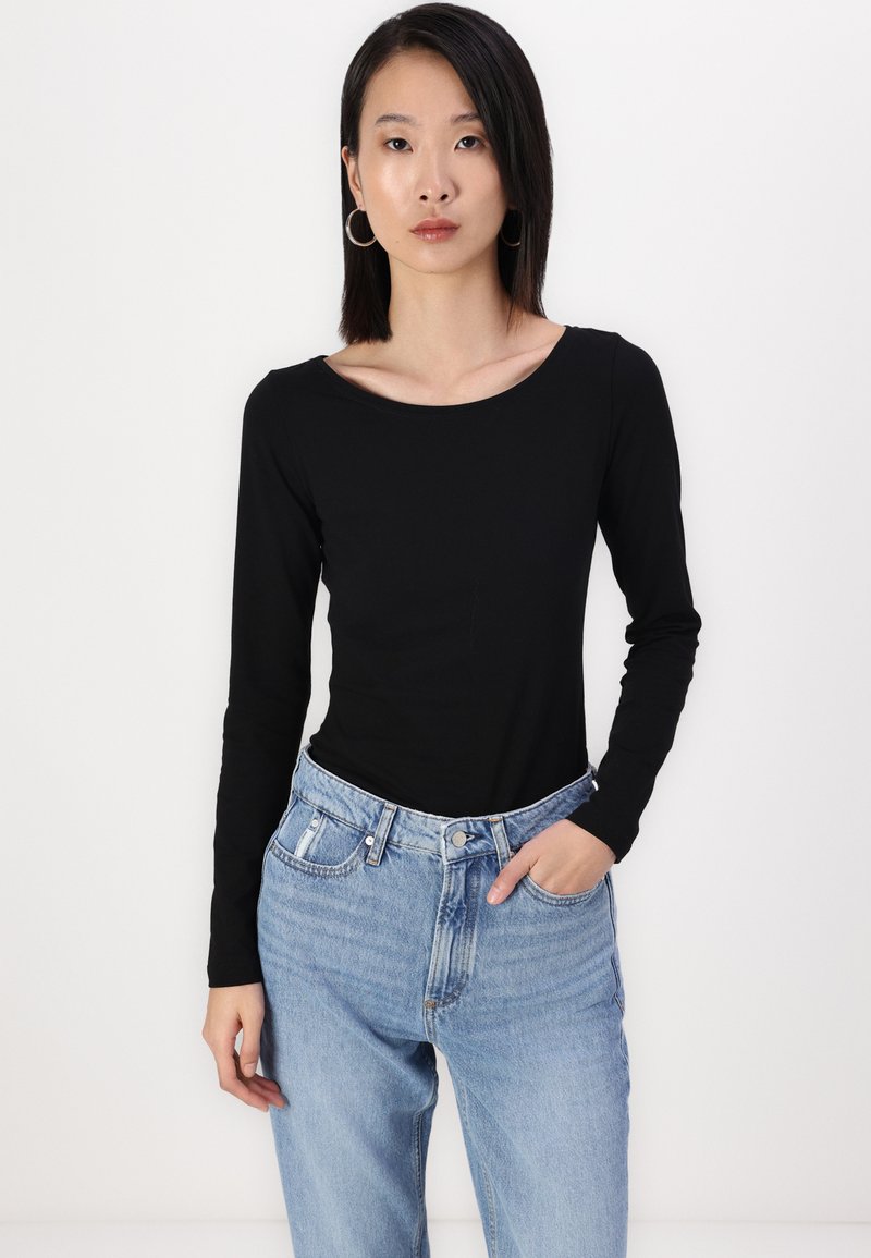 Schwarzes Langarmoberteil mit rundem Halsausschnitt, kombiniert mit hellblauen, hochgeschnittenen Jeans. Das Model steht mit einer Hand in der Tasche vor einem weißen Hintergrund.