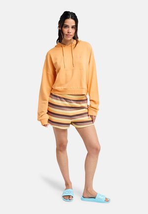 Femme portant un sweat à capuche court orange, un short rayé marron et jaune, et des sandales légères bleu clair, debout devant un fond uni.