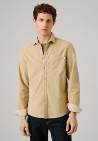 Camicia in velluto a coste beige con taschino sul petto e maniche arrotolate, abbinata a una camicia verde sotto, indossata con jeans scuri.