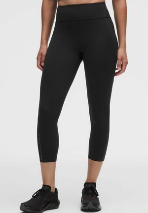 Vrouw draagt zwarte high waist cropped legging met kleine reflecterende stippen op de onderbenen en zwarte sportschoenen, staand op witte achtergrond.