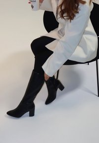 Femme en manteau blanc assise sur une chaise noire, ajustant des bottes noires montantes, tenant une tasse blanche avec du texte.