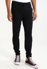 Zwart trainingsbroek met een tapered fit, voorzien van roze zijstrepen en een elastische tailleband. G gedragen met bordeauxrode high-top sneakers.