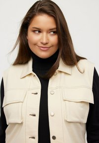 Gilet beige senza maniche con due tasche sul petto, accessori in argento e cuciture in contrasto, indossato sopra un dolcevita nero. Tessuto dalla texture liscia.