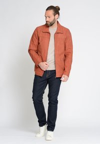 Rostfarbene Wildlederjacke mit Reißverschluss, Stehkragen und seitlichen Taschen, kombiniert mit einem leichten Pullover und dunklen Jeans. Weiße Sneakers.
