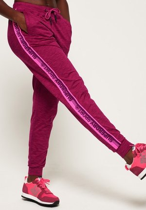 Personne portant un jogging magenta avec des bandes latérales roses et des baskets roses, debout avec une jambe levée sur un fond uni.