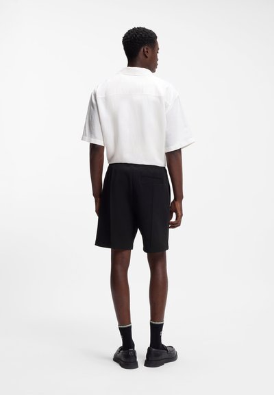Homme portant une chemise blanche ample à manches courtes rentrée dans un short noir arrivant aux genoux, avec des chaussettes noires et des chaussures noires, debout de dos.