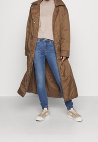 Langer brauner gepolsterter Mantel mit großem Kragen, getragen über einem beigen Pullover und blauen Jeans. Die Schuhe sind beige mit einer weißen Sohle und dem Nike-Logo.