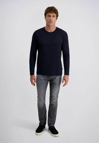Mann steht, trägt ein schwarzes Langarmshirt, ausgewaschene graue Jeans und schwarze Slipper mit weißen Sohlen vor einem schlichten weißen Hintergrund.