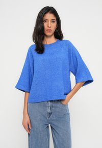 Pull bleu surdimensionné à manches courtes, fabriqué dans un tissu doux et texturé. Associé à un jean en denim clair, présentant une coupe décontractée.
